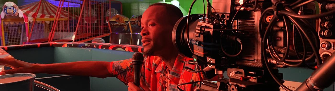 African American man holding microphone. Camera in right foreground pointing at him.